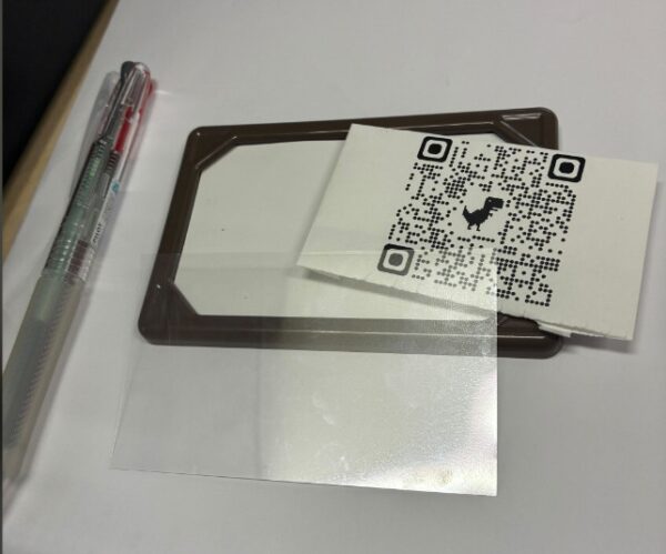 ネームプレート、QRコードが印刷された紙、プラスチックのカバーが写っている。またサイズの比較用にペンがおかれている。